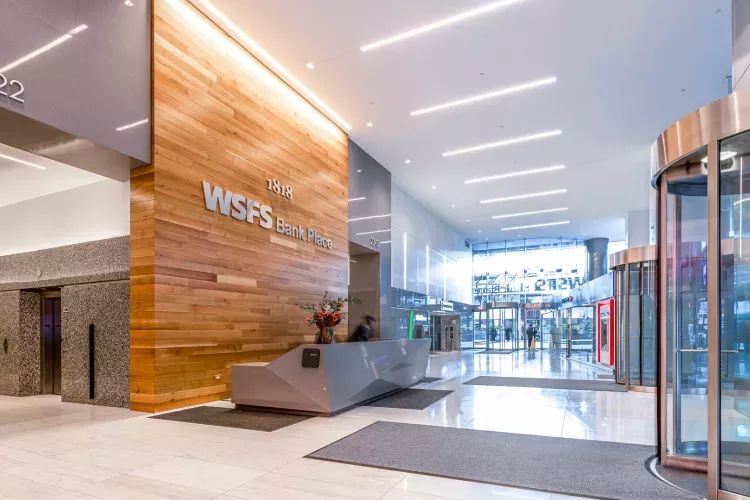 Spacious and modern lobby of WSFS Bank Place at 1818 Market, featuring a wood-paneled accent wall with building signage, polished marble floors, sleek revolving glass doors, granite elevator banks, and a geometric reception desk under soft overhead lighting.