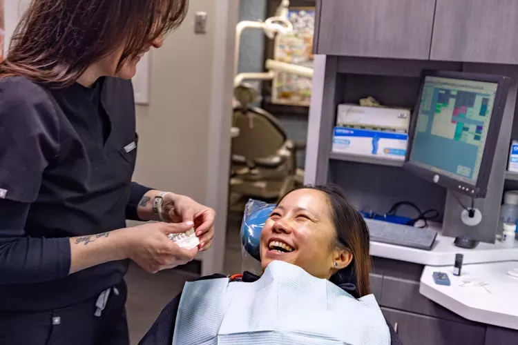 A smiling patient sits in a dental chair while a dental professional shows a model of teeth, creating a warm and welcoming atmosphere in a modern dental office with a computer workstation and dental equipment visible in the background.